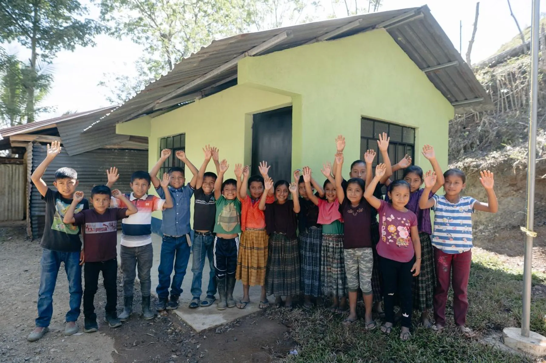 Niñas y niños saludando frente a un aula construida en su comunidad